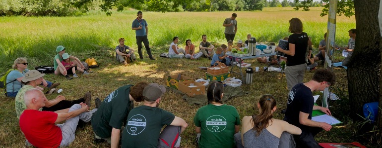 Dobrovolníci sedí v kruhu na louce a poslouchají výklad lektora.